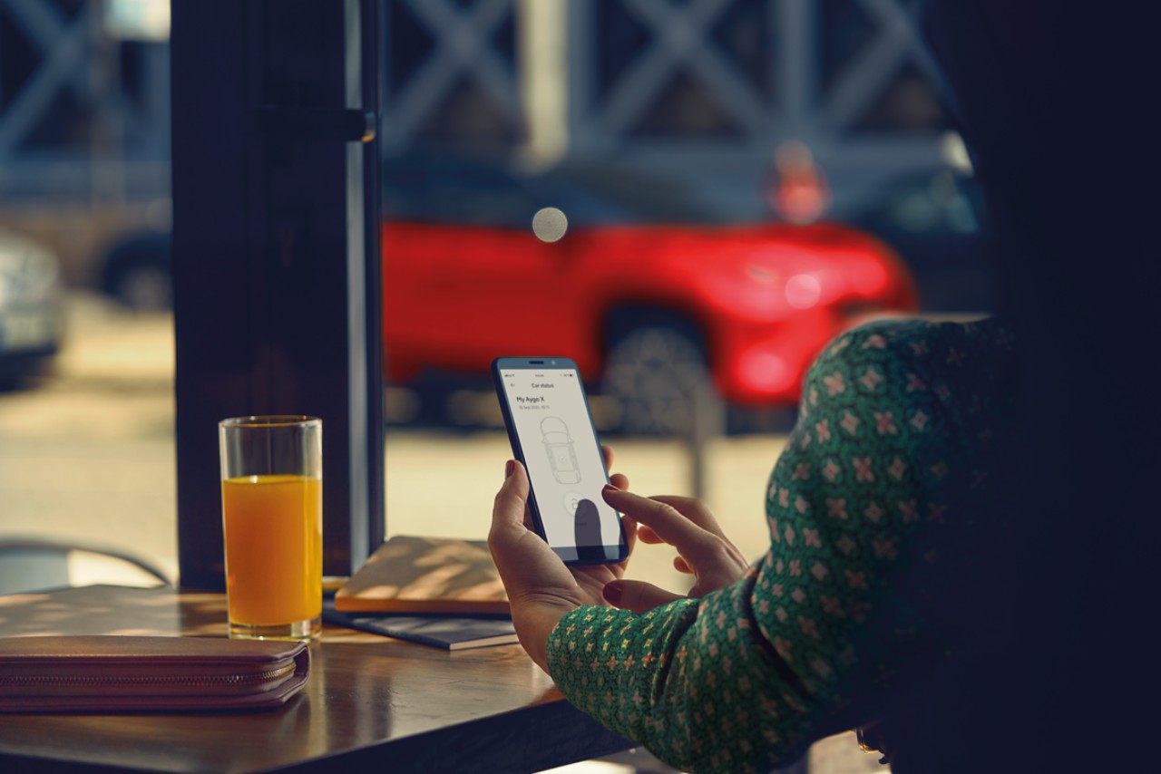 Eine Frau sitzt an einem Restauranttisch und benutzt die MyToyota-App auf ihrem Telefon, ihr Toyota Yaris Cross ist im Hintergrund geparkt.