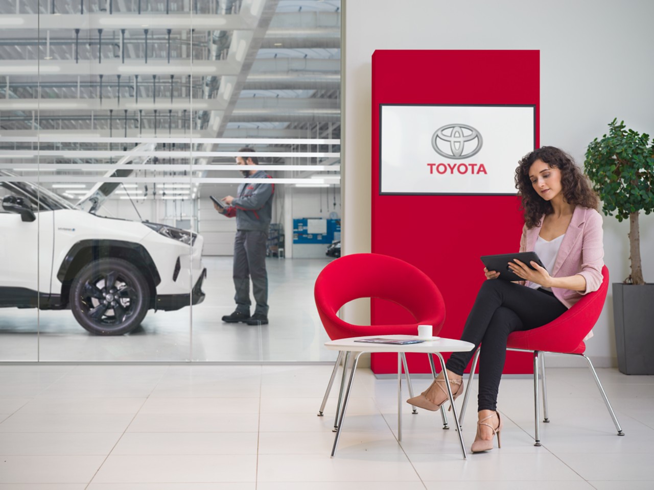 Eine Frau wartet in einem Toyota Servicebetrieb und schaut auf ihr Tablet. Hinter ihr ein Bildschirm mit Toyota Logo.