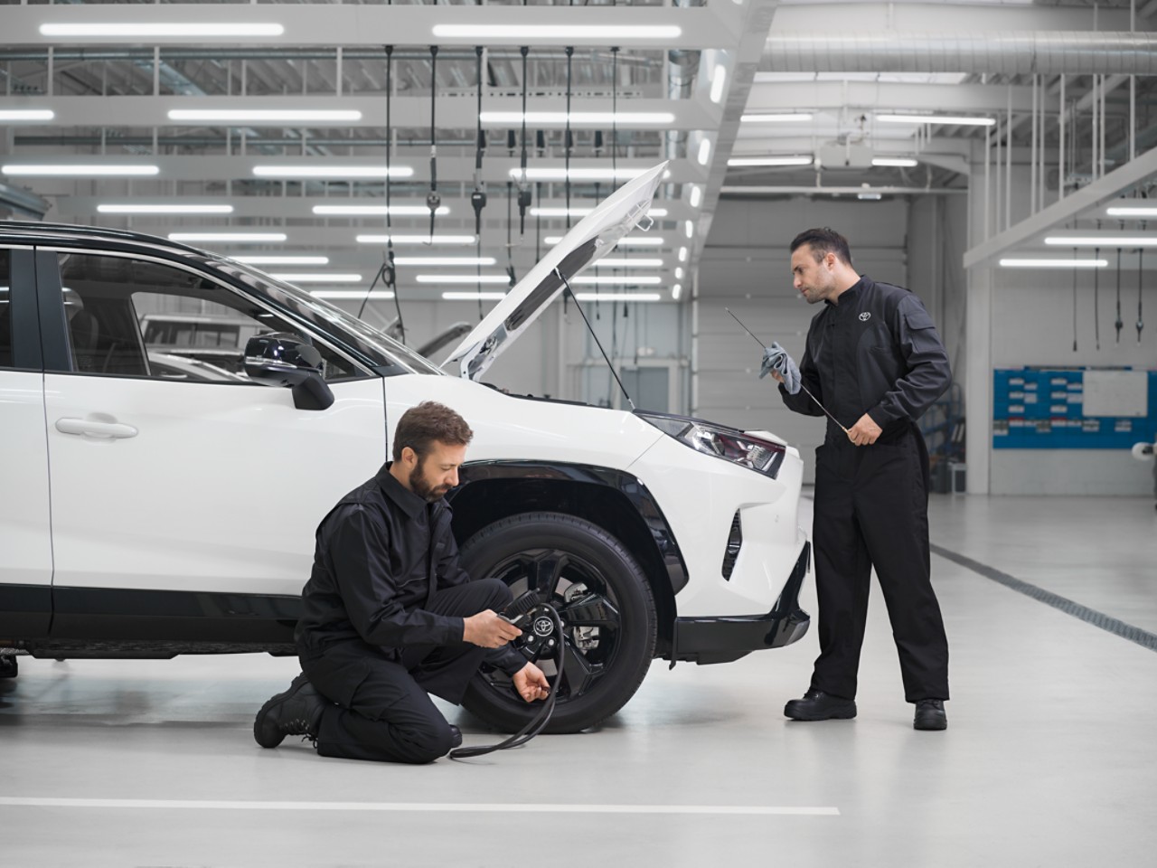 Zwei Techniker arbeiten an einem weissen Toyota RAV-4 mit offener Motorhaube. Einer kontrolliert den Reifendruck, der andere den Ölstand.
