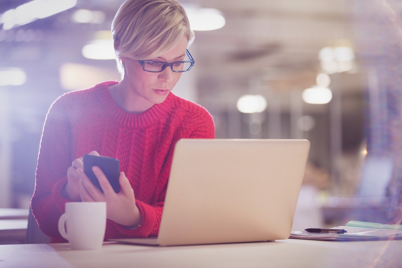 Eine Frau mit Brille und rotem Pullover, in der Hand ein Smartphone, schaut auf ihren Laptop. Auf dem Tisch: eine Kaffeetasse. 
