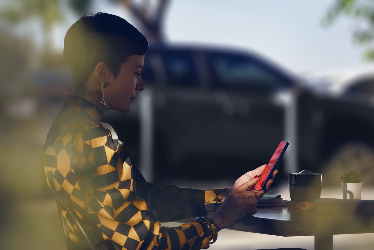 Eine Frau sitzt an einem Restauranttisch und benutzt die MyToyota-App auf ihrem Telefon, ihr Toyota Yaris Cross ist im Hintergrund geparkt.